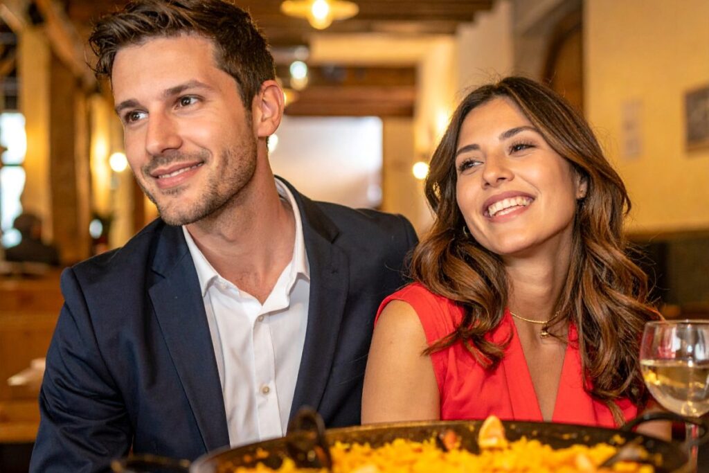 en el restaurante - Prueba Esto Hoy Un hombre y una mujer jóvenes sonriendo, sentados en un restaurante delante de una paella.