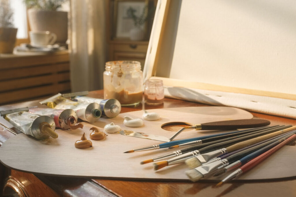 Materiales de pintura sobre mesa de estudio: tubos de óleo y acrílico abiertos, pinceles de cerdas duras y sintéticas, espátula, pequeñas manchas de pintura espesa sobre una paleta, lienzo preparado y frascos de médium. Imagen sintética.