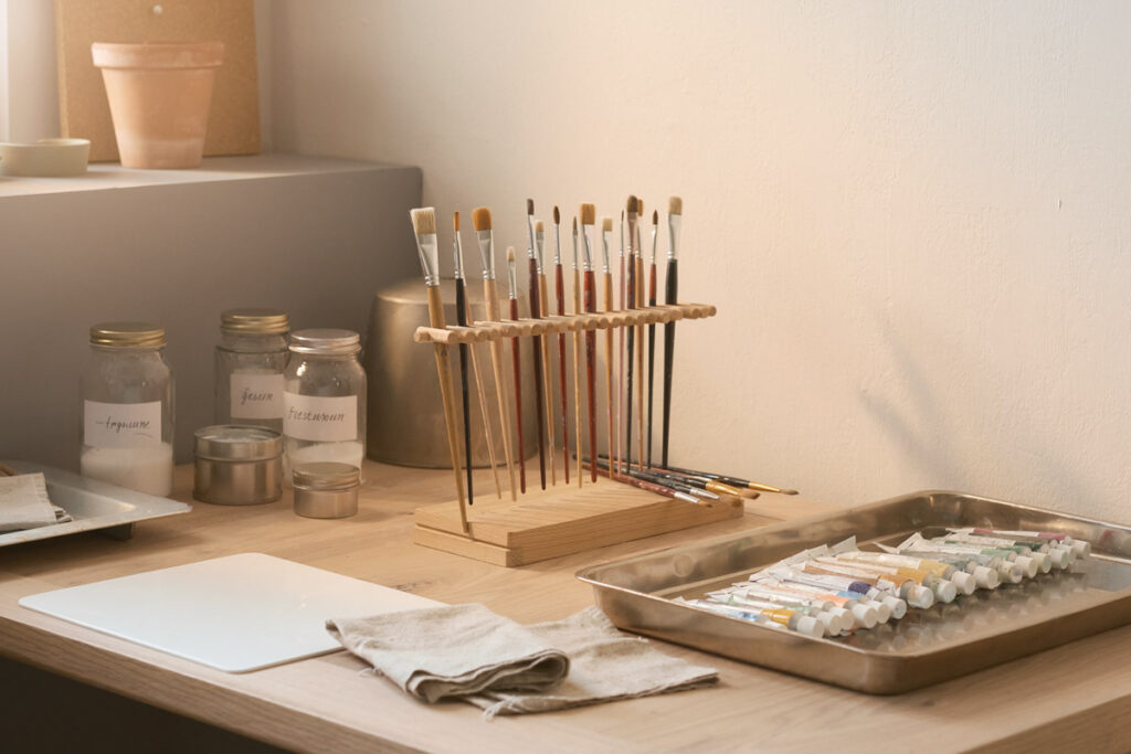 Un rincón de estudio de arte ordenado, con pinceles limpios secándose en horizontal, tubos de pintura bien cerrados, recipientes etiquetados, paños, paleta limpia y materiales organizados en bandejas. Estética minimalista, sensación de hábito consciente y cuidado de herramientas. Imagen sintética.
