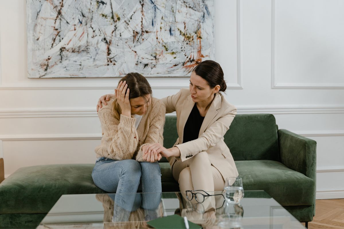 Una mujer consolando a una amiga en su casa. Ambas están sentadas enun sofá.