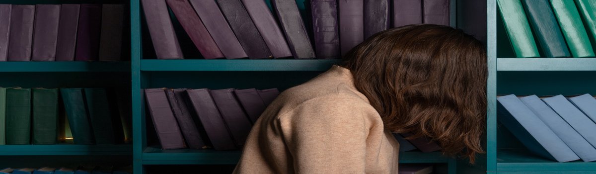 mujer triste - Prueba Esto Hoy La imagen muestra a una mujer joven triste con libros en estanterias como fondo.