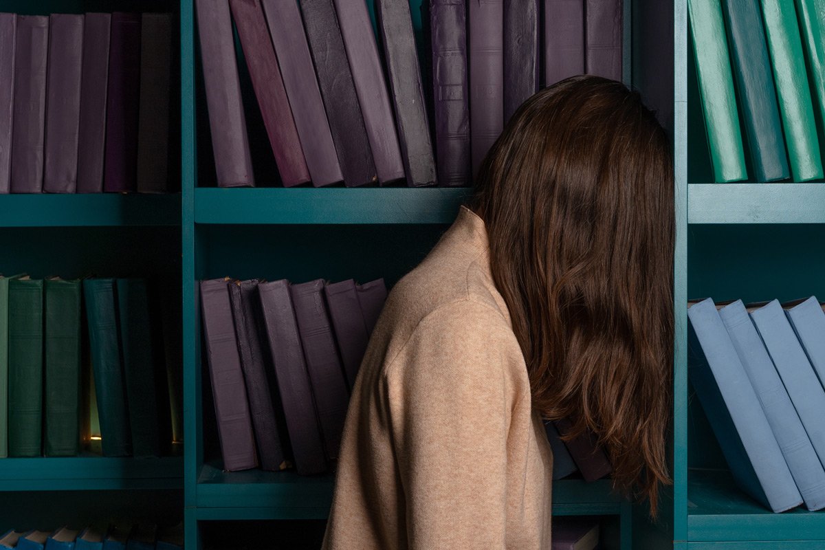 La imagen muestra a una mujer joven triste con libros en estanterias como fondo.