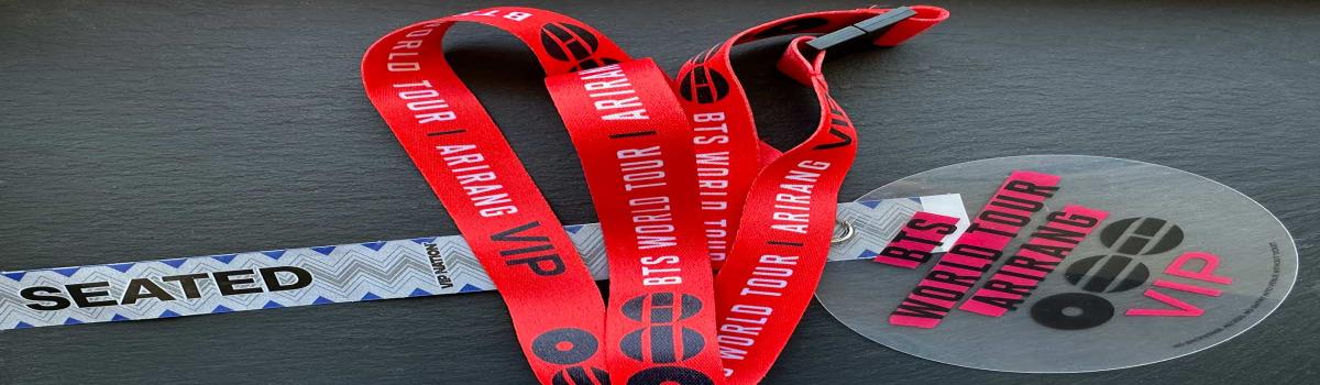Cintas VIP rojas con los logotipos de «WORLD TOUR ARIRANG» y BTS de las cuales cuelga una acreditación VIP redonda de color gris y la pulsera para la prueba de sonido junto a ellas.