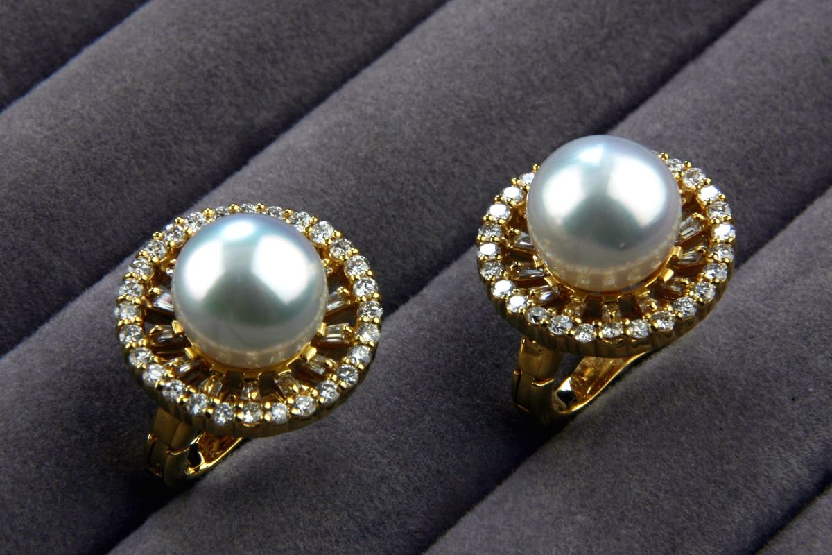 Two gold rings each featuring a large white pearl surrounded by a circle of diamonds on a dark fabric background, shown from above.