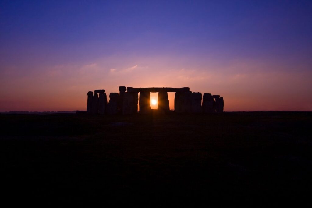 Silueta de Stonehenge al atardecer, con el sol brillando a través de la abertura central sobre un cielo de tonos violeta y naranja.
