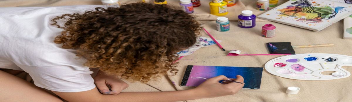 Niña pintando con acrílicos sobre un pequeño lienzo, rodeada de pinceles, una paleta y botes de pintura de varios colores.