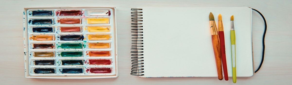 Una caja de acuarelas, un cuaderno para pintar y unos pinceles encima del cuaderno.