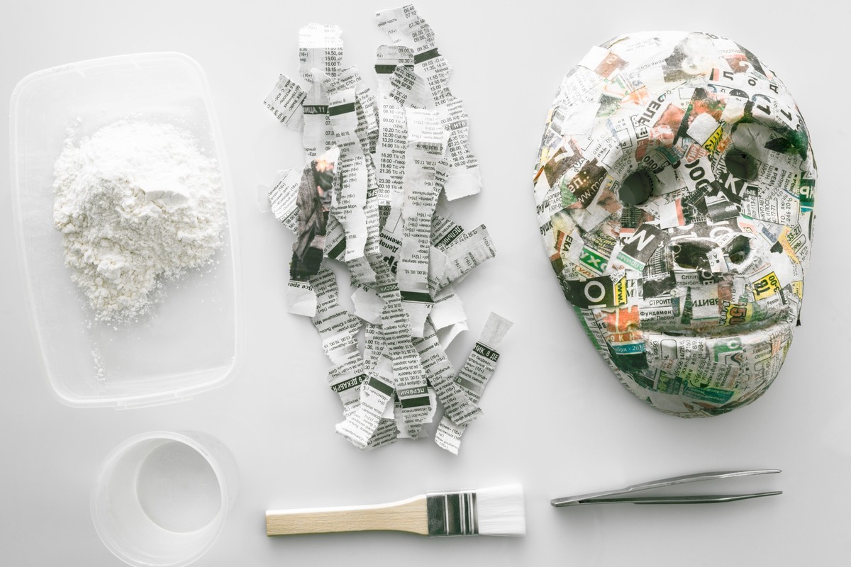 Materiales para hacer papel maché dispuestos sobre una superficie blanca: un recipiente con engrudo de harina, tiras de periódico rasgadas, una máscara de periódico terminada, un cuenco con agua, un pincel y unas pinzas