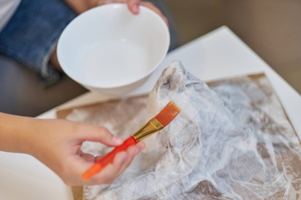 Un niño aplica engrudo con un pincel sobre una forma cubierta de plástico en una bandeja forrada, dando forma a una montaña de papel maché, con un cuenco blanco sostenido encima.